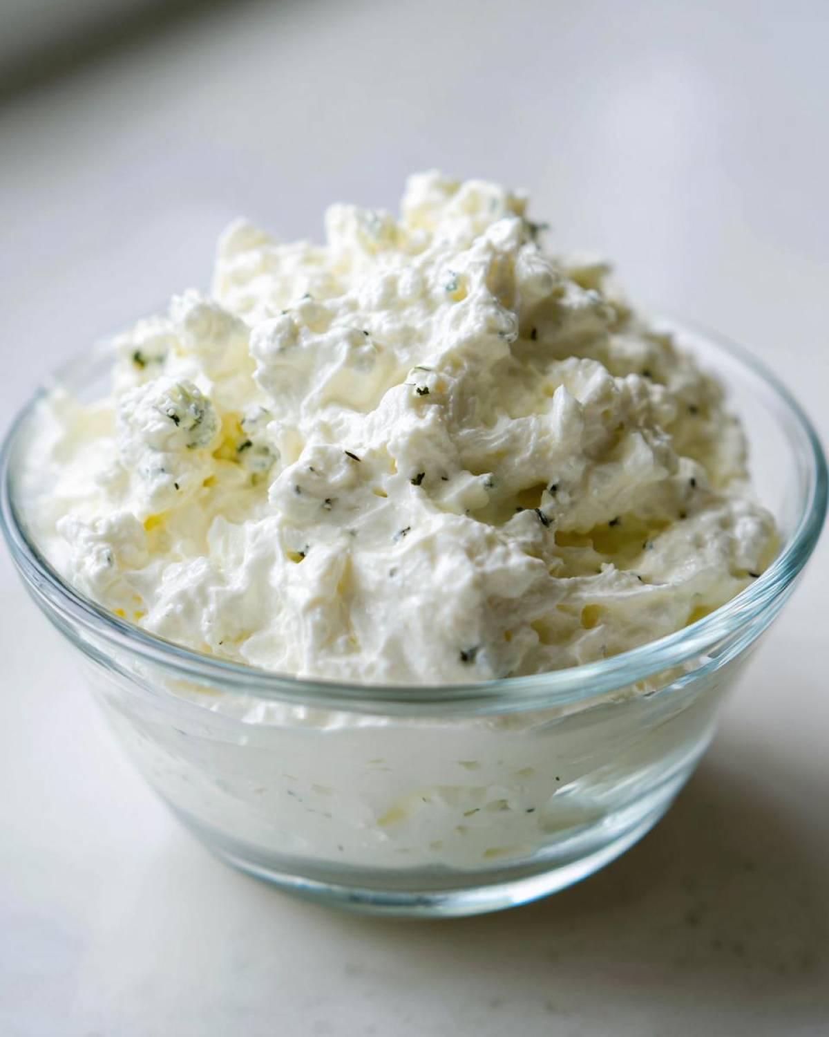 Close-up of a fluffy, white blue cheese dip with visible blue cheese crumbles and herbs in a clear glass bowl.