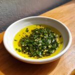 Close-up of a white bowl containing vibrant olive oil bread dip with chopped green herbs and garlic.