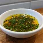 Close-up of a small white bowl filled with golden olive oil bread dip topped with chopped green herbs and garlic.
