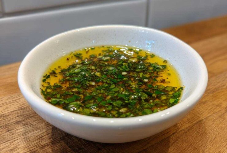 Close-up of a small white bowl filled with golden olive oil bread dip topped with chopped green herbs and garlic.