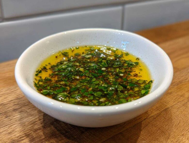 Close-up of a small white bowl filled with golden olive oil bread dip topped with chopped green herbs and garlic.