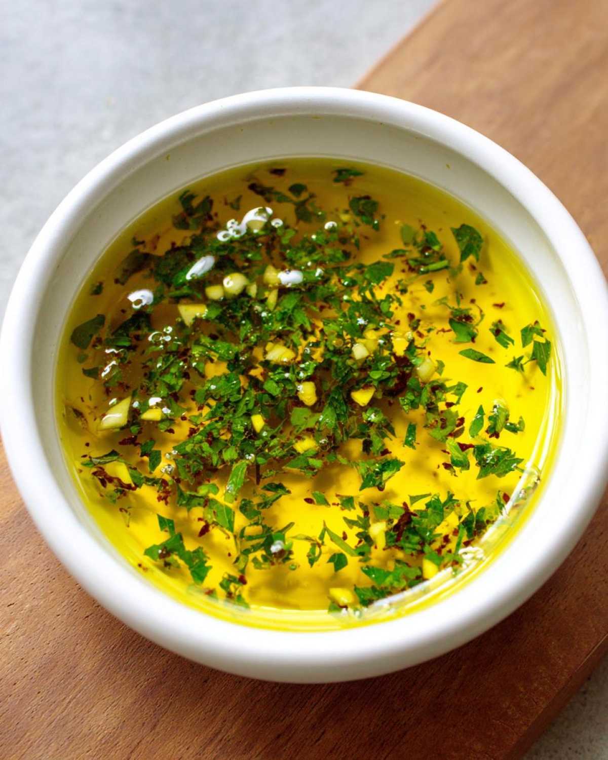 Close-up of a white bowl filled with vibrant yellow olive oil bread dip, topped with fresh green herbs and minced garlic.