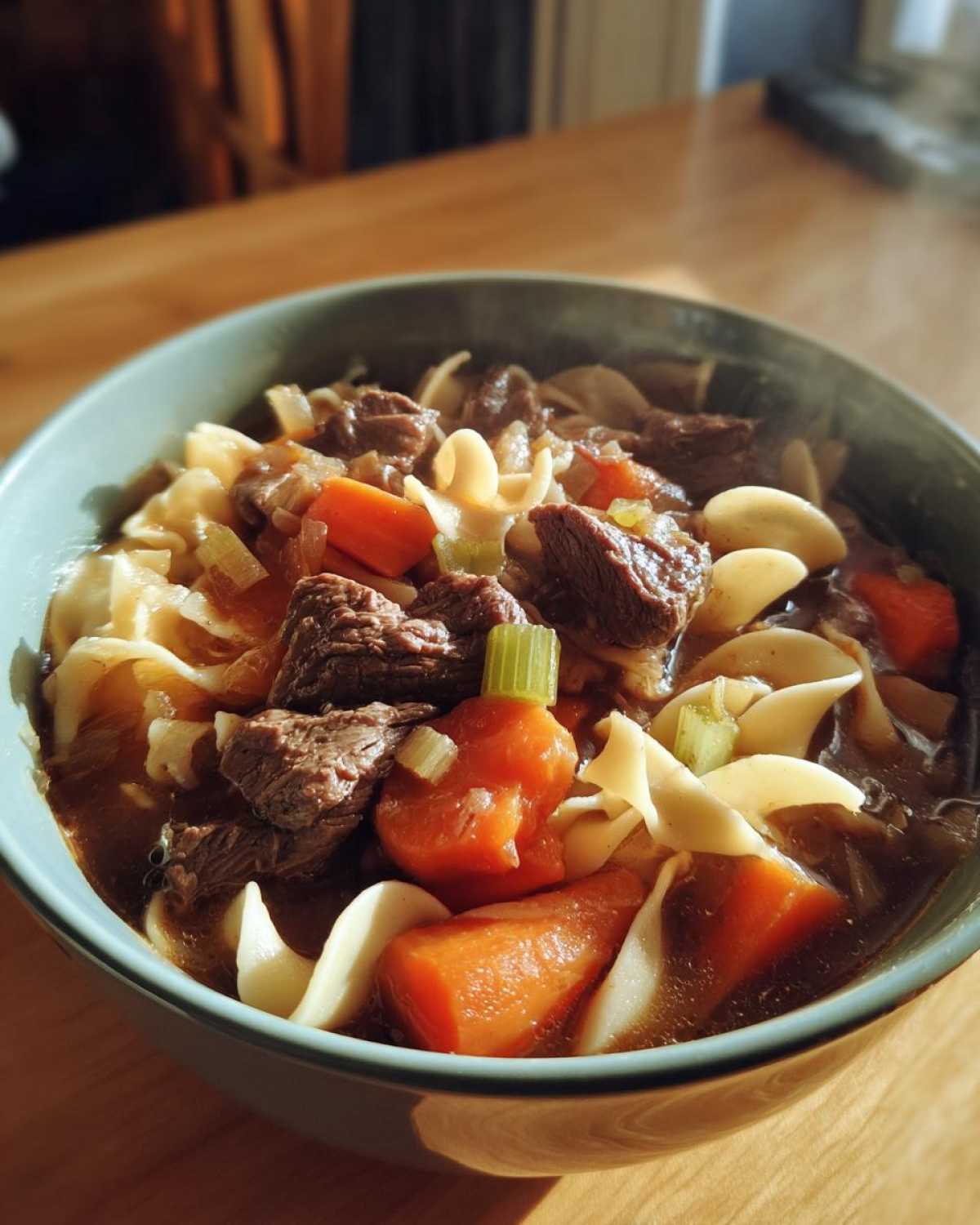 Close-up of a steaming bowl of amazing beef noodle soup with chunks of beef, carrots, and egg noodles.