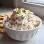 A white bowl filled with creamy clam dip, topped with green onions, next to a stack of crackers.