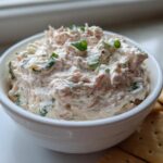 A creamy, pinkish-white clam dip topped with green onions, served in a white bowl next to crackers.