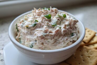 A creamy, pinkish-white clam dip topped with green onions, served in a white bowl next to crackers.