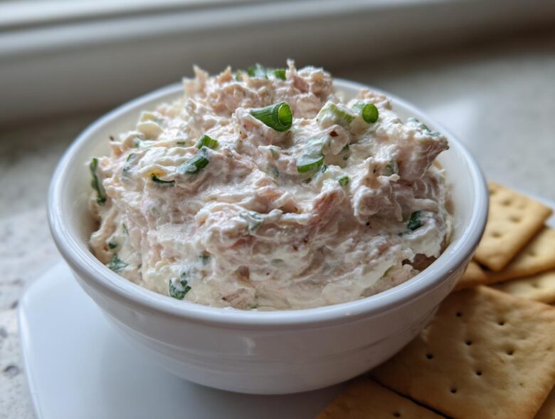 A creamy, pinkish-white clam dip topped with green onions, served in a white bowl next to crackers.