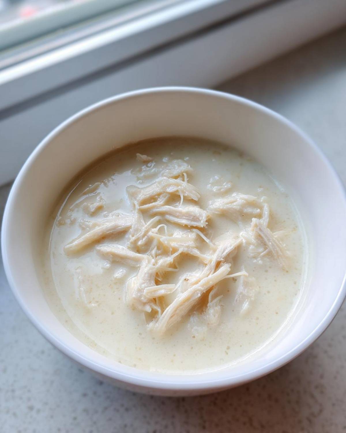 A close-up of a white bowl filled with creamy, homemade cream of chicken soup topped with shredded white meat chicken.