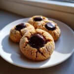 Three freshly baked kiss cookies, rolled in sugar and topped with melted chocolate, sitting on a white plate.