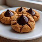 Three freshly baked peanut butter kiss cookies, rolled in sugar and topped with a chocolate candy, sitting on a white plate.