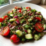 A close-up of a fresh lentil salad featuring brown lentils, chopped cucumber, halved cherry tomatoes, and red onion in a white bowl.