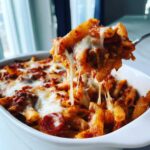 A fork lifting a serving of baked pizza pasta, showing extreme cheese pull over a casserole dish.
