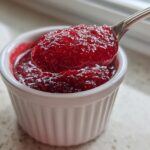 Close-up of a spoonful of thick, bright red raspberry jam being lifted from a small white ramekin.