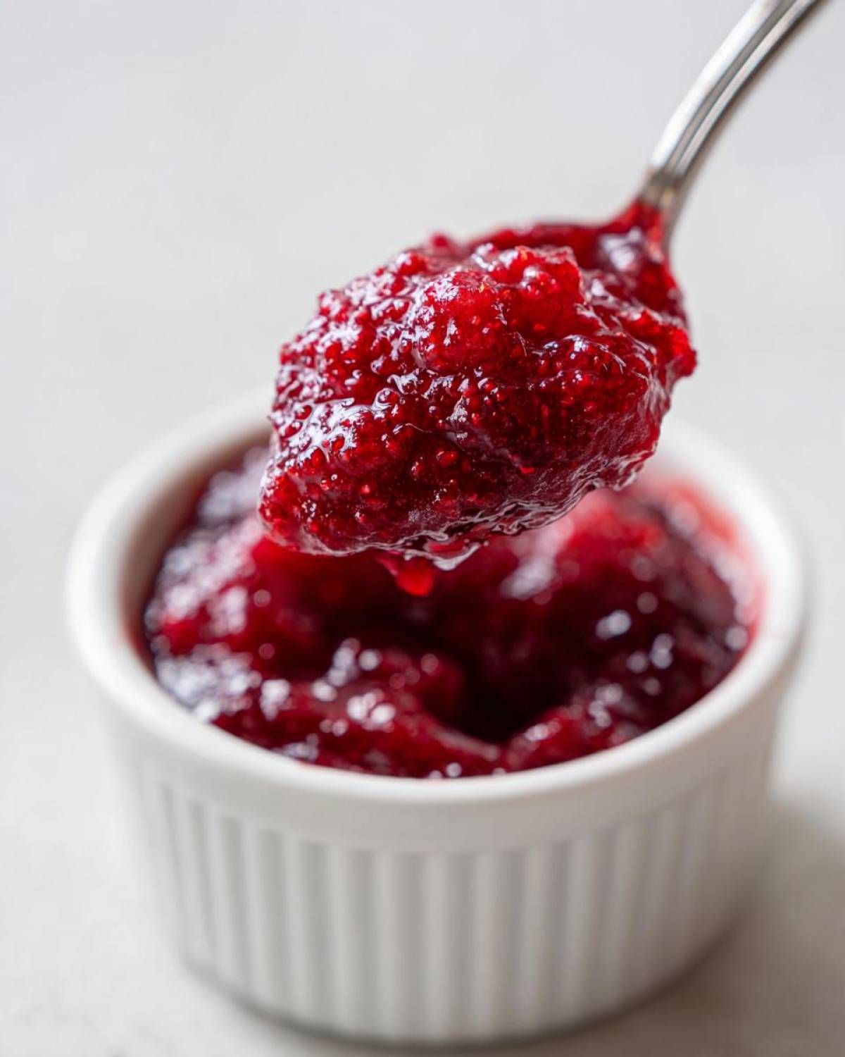 A spoonful of thick, vibrant red homemade raspberry jam being lifted above a small white ramekin.