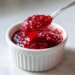A spoonful of glistening, seed-filled homemade raspberry jam being lifted from a small white ramekin.