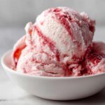 Close-up of creamy scoops of homemade strawberry ice cream with visible red fruit swirls in a white bowl.