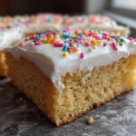 A close-up of a square, thick sugar cookie bars slice topped with white frosting and colorful sprinkles.