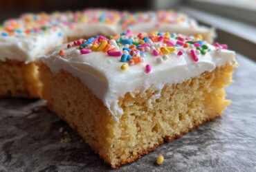 A close-up of a square, thick sugar cookie bars slice topped with white frosting and colorful sprinkles.