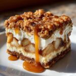 A thick, layered slice of apple cheesecake bars topped with crumble and dripping with caramel sauce.