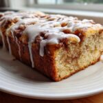 A freshly baked loaf of apple fritter bread drizzled heavily with white icing, sitting on a white plate.