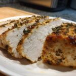 Close-up of juicy, sliced baked chicken breast seasoned with herbs on a white plate.