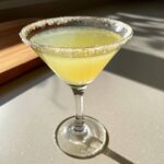 A bright yellow lemon drop cocktail served in a martini glass with a sugar-rim, catching bright sunlight.