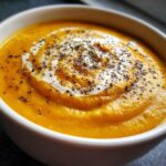 Close-up of a bowl of vibrant orange butternut squash soup, swirled with cream and topped with cracked black pepper.