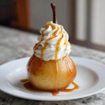 A single, glossy poached pear topped with a swirl of whipped cream and caramel sauce.
