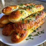 A stack of golden brown, cheesy garlic breadsticks topped with chopped fresh parsley on a white plate.