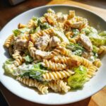 A close-up, appetizing shot of chicken caesar pasta salad featuring fusilli pasta, chicken chunks, romaine lettuce, and croutons.