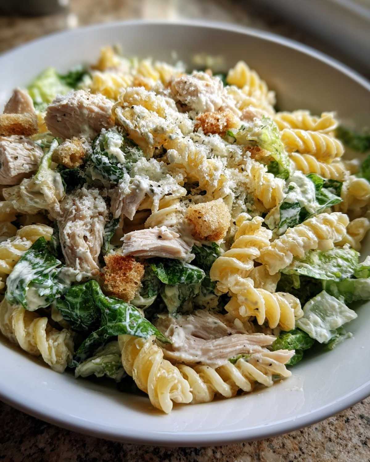 A close-up view of a white bowl filled with chicken caesar pasta salad, featuring rotini pasta, chicken pieces, romaine lettuce, and grated cheese.