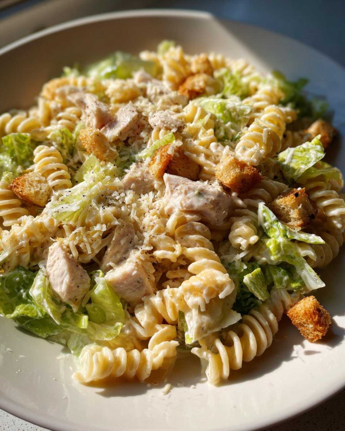 A close-up shot of a creamy chicken caesar pasta salad featuring fusilli pasta, diced chicken, romaine lettuce, and croutons.