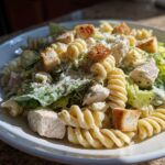 Close-up of a white plate filled with creamy chicken caesar pasta salad, featuring rotini pasta, chicken chunks, romaine lettuce, and croutons.