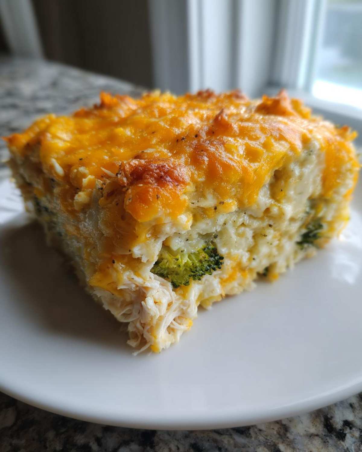 A square serving of cheesy chicken cauliflower rice casserole showing shredded chicken and broccoli florets.