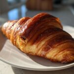 Close-up of a flaky, golden-brown chocolate croissant resting on a white plate in bright sunlight.