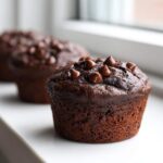 Close-up of a rich, dark chocolate zucchini muffin topped with melted chocolate chips, sitting on a white surface.