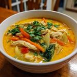A close-up shot of a steaming bowl of rich, yellow coconut curry soup loaded with shredded chicken, carrots, bok choy, and fresh cilantro.