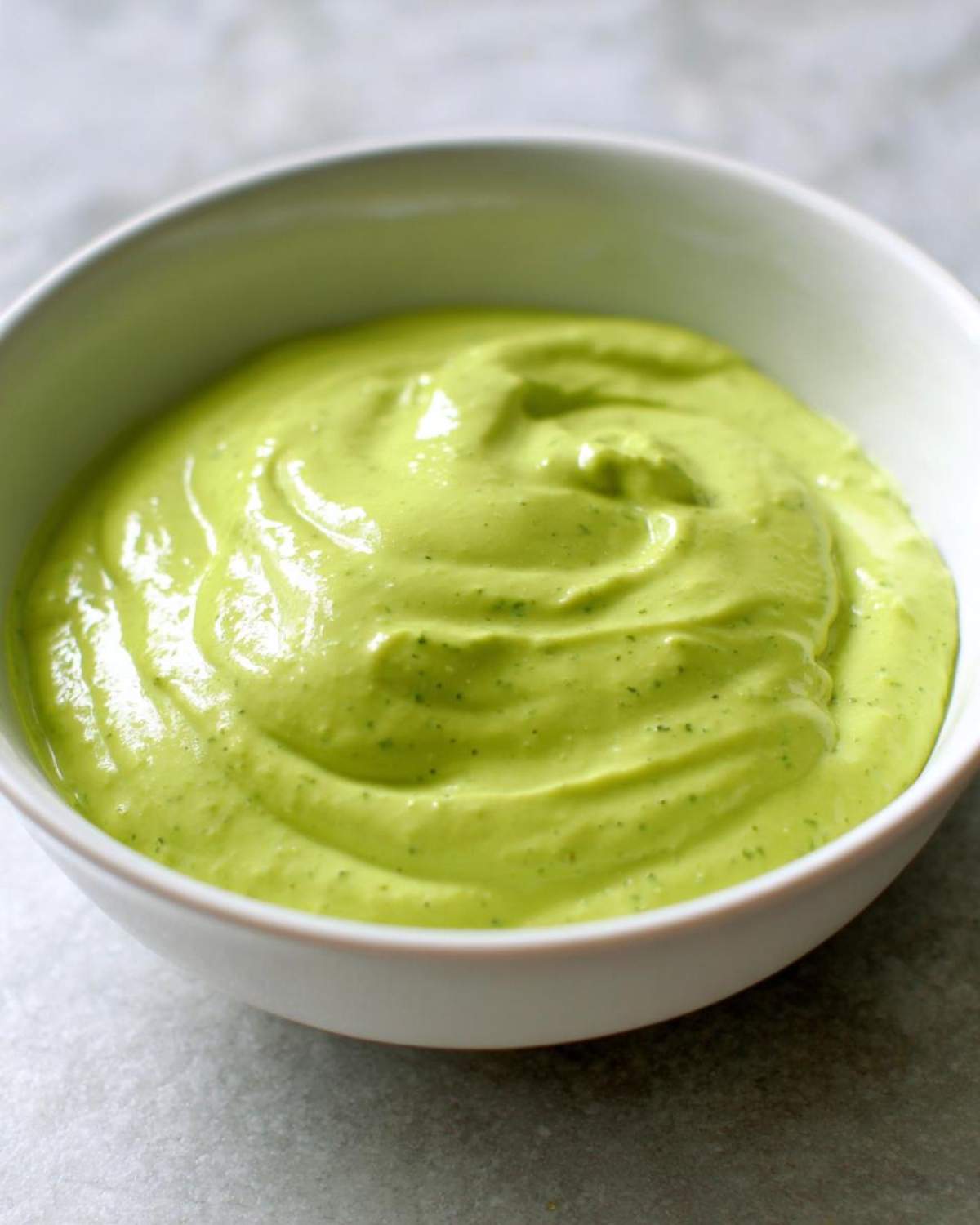 Close-up shot of thick, vibrant green avocado sauce swirled in a small white bowl.