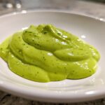 A close-up of a thick, vibrant green dollop of creamy avocado sauce speckled with herbs, served in a small white bowl.