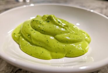 A close-up of a thick, vibrant green dollop of creamy avocado sauce speckled with herbs, served in a small white bowl.
