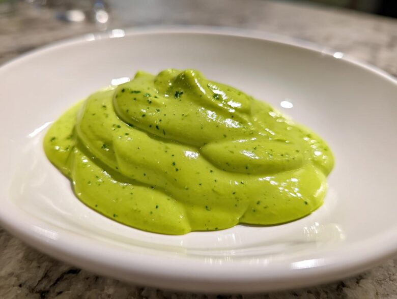 A close-up of a thick, vibrant green dollop of creamy avocado sauce speckled with herbs, served in a small white bowl.