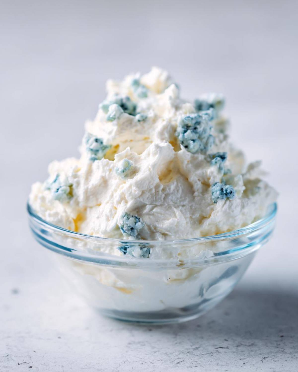 A close-up of a fluffy, creamy blue cheese dip garnished with visible blue cheese crumbles in a clear glass bowl.