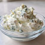 A close-up view of thick, creamy blue cheese dip with visible blue mold chunks served in a small clear glass bowl.