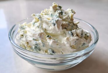 A close-up view of thick, creamy blue cheese dip with visible blue mold chunks served in a small clear glass bowl.
