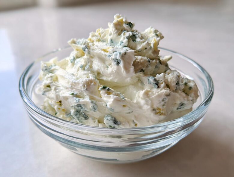 A close-up view of thick, creamy blue cheese dip with visible blue mold chunks served in a small clear glass bowl.