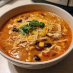 A close-up of a white bowl filled with creamy chicken enchilada soup, topped with shredded chicken, cheese, corn, and cilantro.
