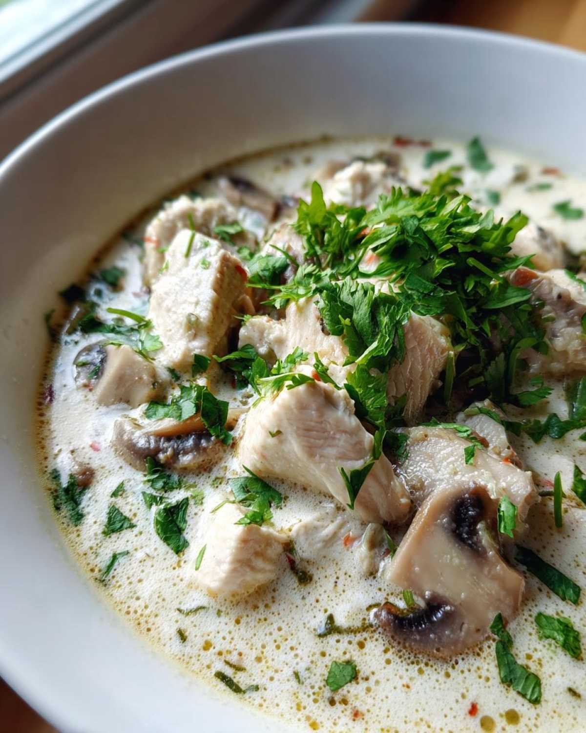 Close-up of a bowl of creamy coconut soup featuring chunks of chicken, sliced mushrooms, and fresh parsley garnish.