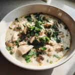 A close-up of a white bowl filled with creamy chicken and mushroom coconut soup, topped generously with fresh cilantro.