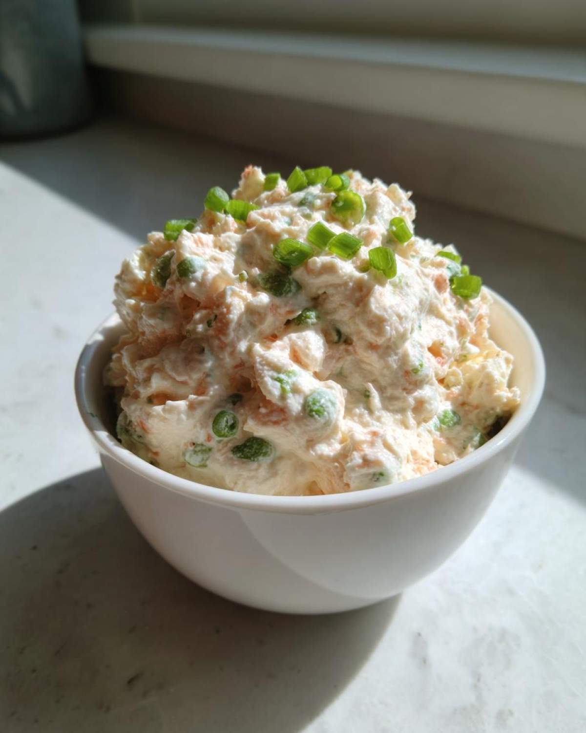 A close-up of a bowl of creamy clam dip mixed with orange shreds and green peas, topped with chopped chives.