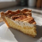 A close-up of a thick slice of creamy custard pie topped with meringue and dusted heavily with cinnamon.